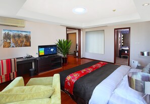 Una camera d'albergo con un letto grande, una scrivania con un computer, una TV, una pianta e un bagno visibile attraverso una porta aperta.