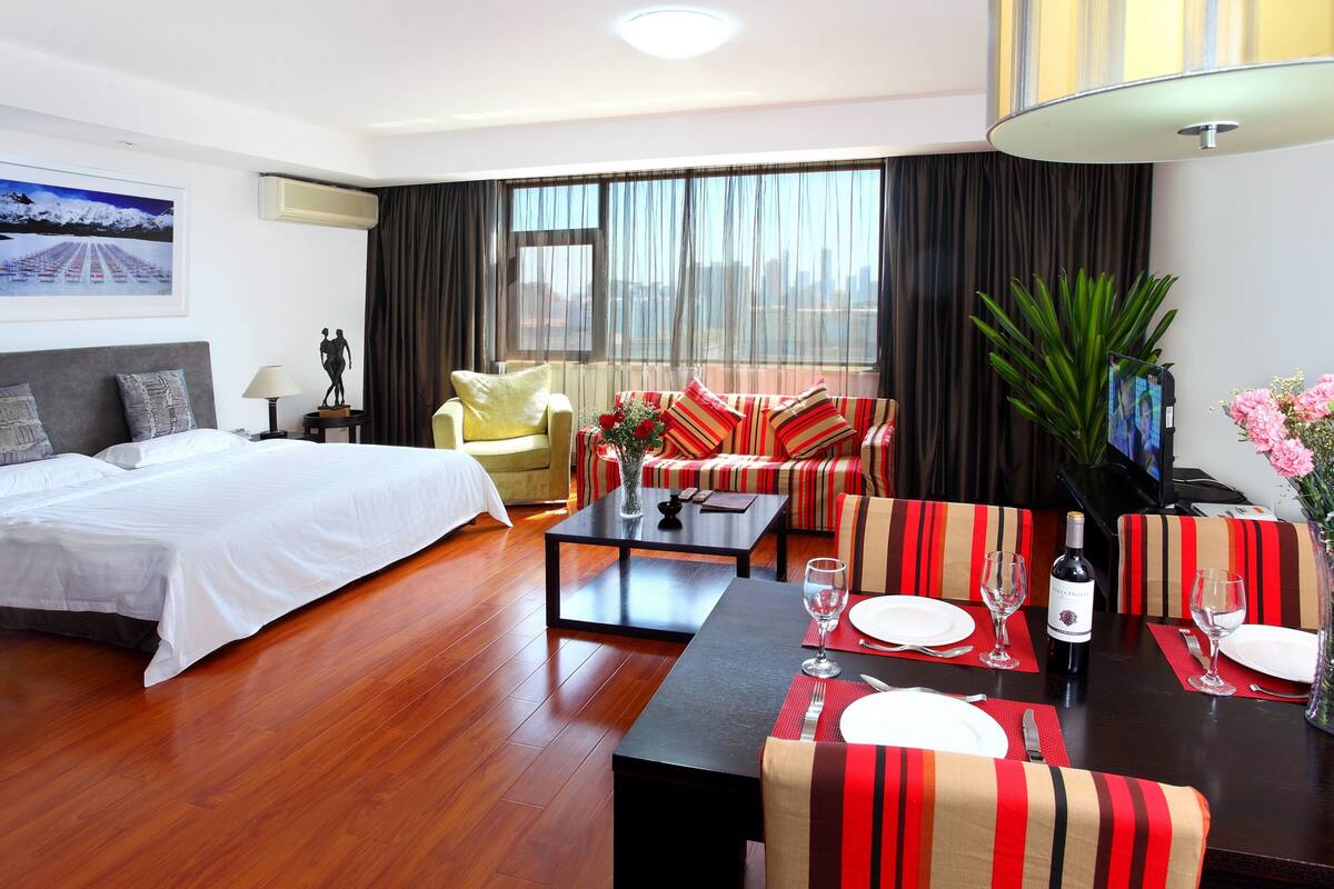 Quarto de hotel com cama, sofá, mesa de jantar posta para dois e uma janela grande com cortinas.