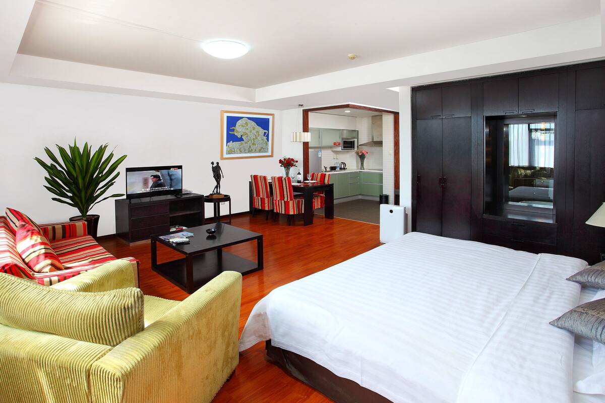 Quarto de hotel moderno com cama grande, sofá, mesa de jantar e cozinha visível por uma porta aberta.