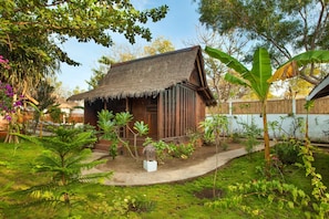 A rustic wooden house with a thatched roof, surrounded by lush greenery and a variety of plants.