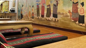 Ein Zimmer mit einer Wandmalerei, die traditionelle Figuren zeigt, einem an der Wand montierten Flachbildfernseher und einem Holztisch mit Kissen.