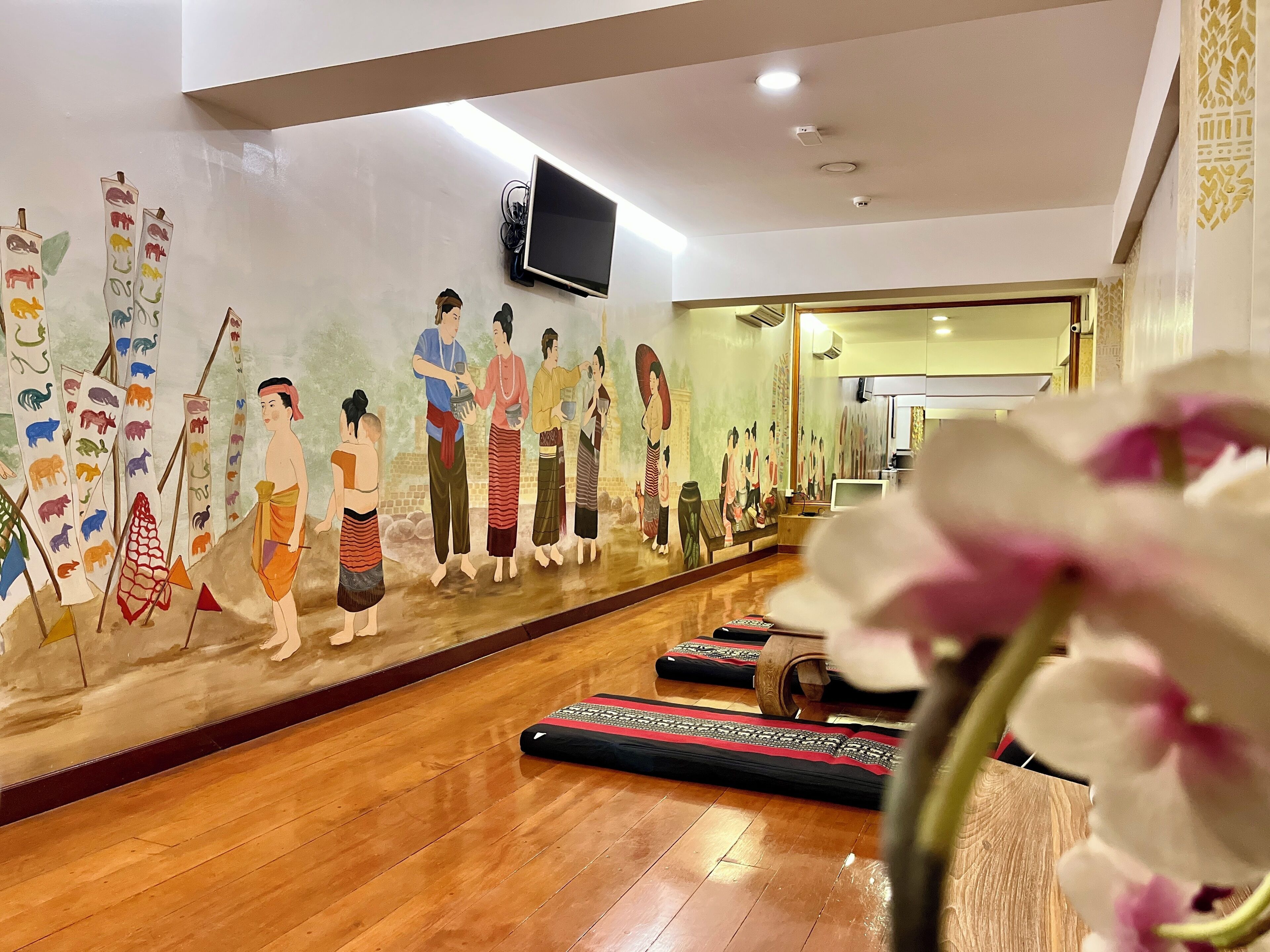 A room with a mural depicting traditional figures, wooden flooring, and a television mounted on the wall.