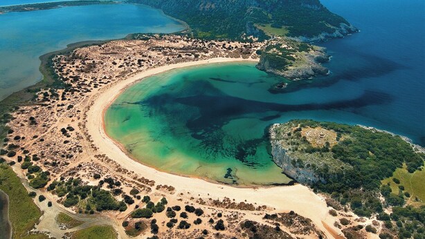 Eine Luftaufnahme einer Küstenregion mit Sandstrand, klarem Wasser und einem Bergpanorama im Hintergrund.