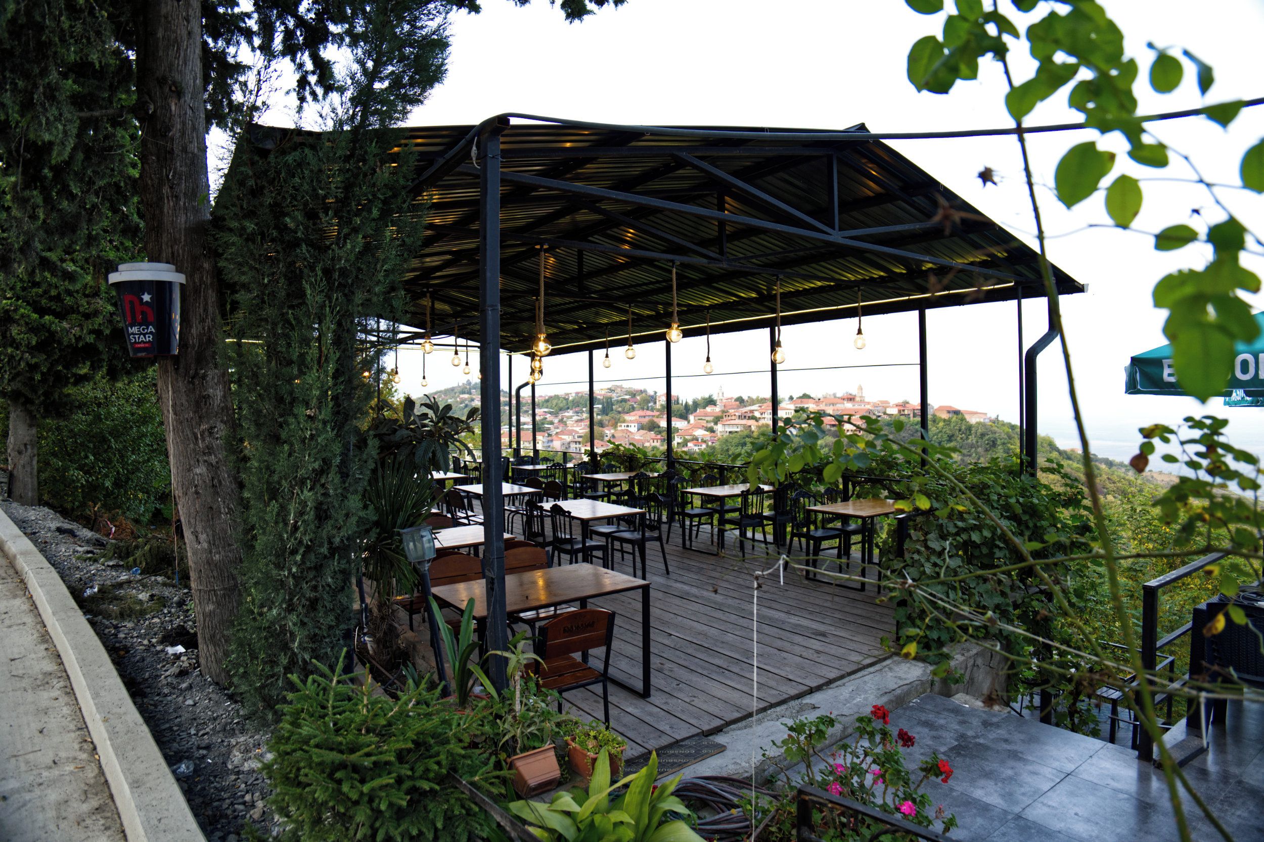 An outdoor seating area with tables and chairs, surrounded by greenery and overlooking a cityscape.