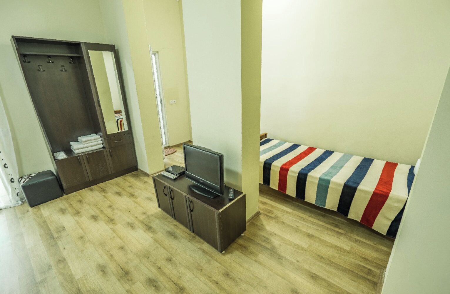 A room with a wooden floor, a bed with a striped blanket, a TV on a cabinet, and a wardrobe.