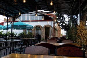 An outdoor cafe with tables and chairs, a brick building with arched windows, and hanging light bulbs.