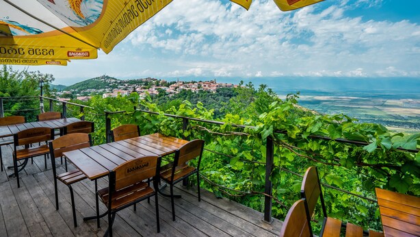 Un espace extérieur aménagé avec des tables et des chaises en bois, entouré d’une végétation luxuriante et offrant une vue sur une ville lointaine.