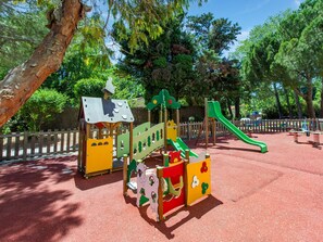Children can play on a slide, climbing frames, and a playhouse in this fenced play area surrounded by trees.