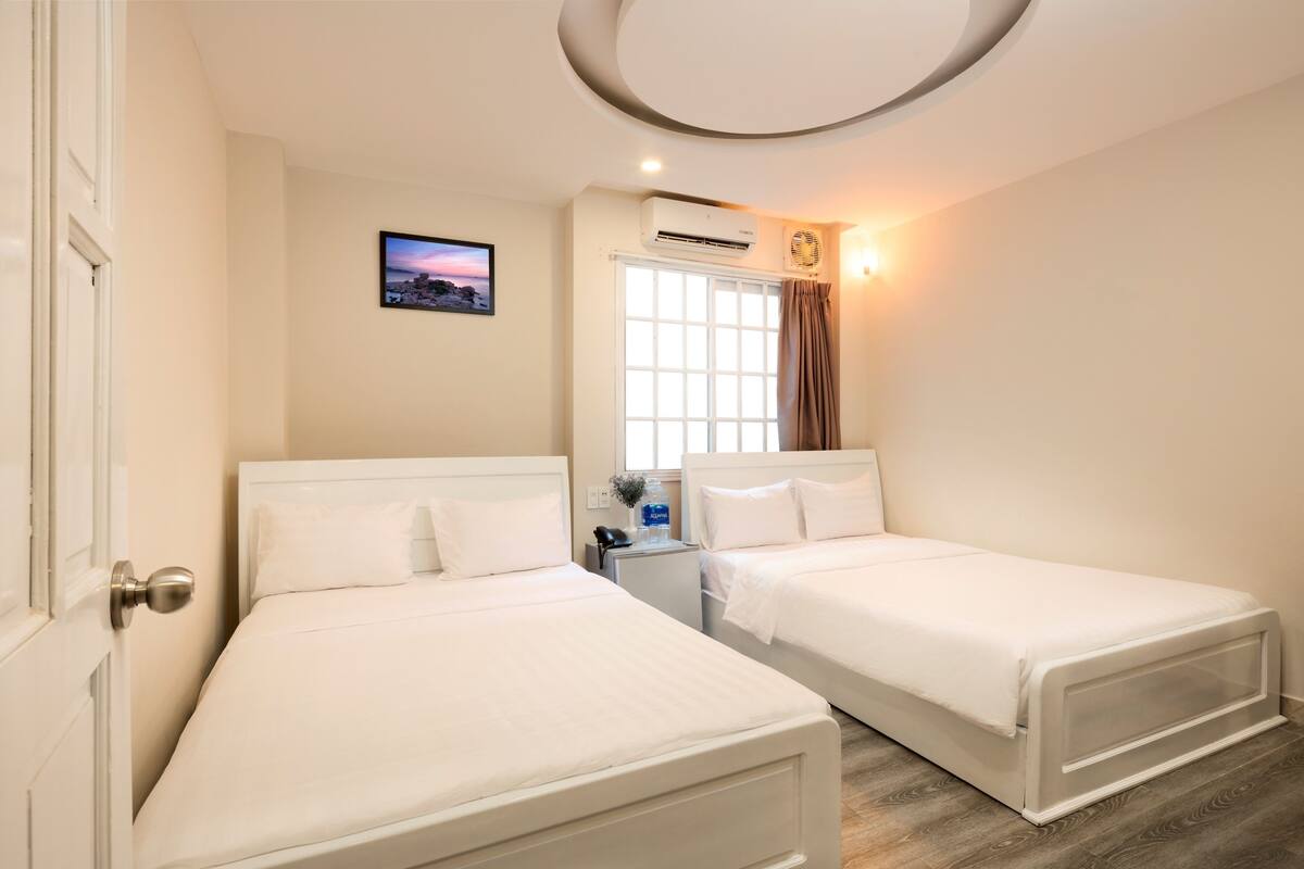 Quarto de hotel com duas camas de solteiro, janela com cortinas, ar condicionado, mesa pequena com telefone e um vaso, e um quadro na parede.