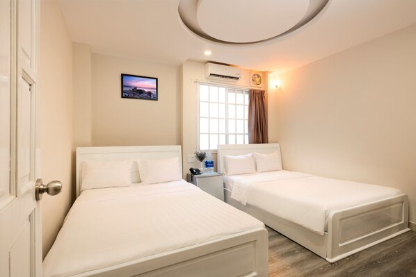 A hotel room with two single beds, a window with curtains, an air conditioning unit, a small table with a phone and a vase, and a picture on the wall.
