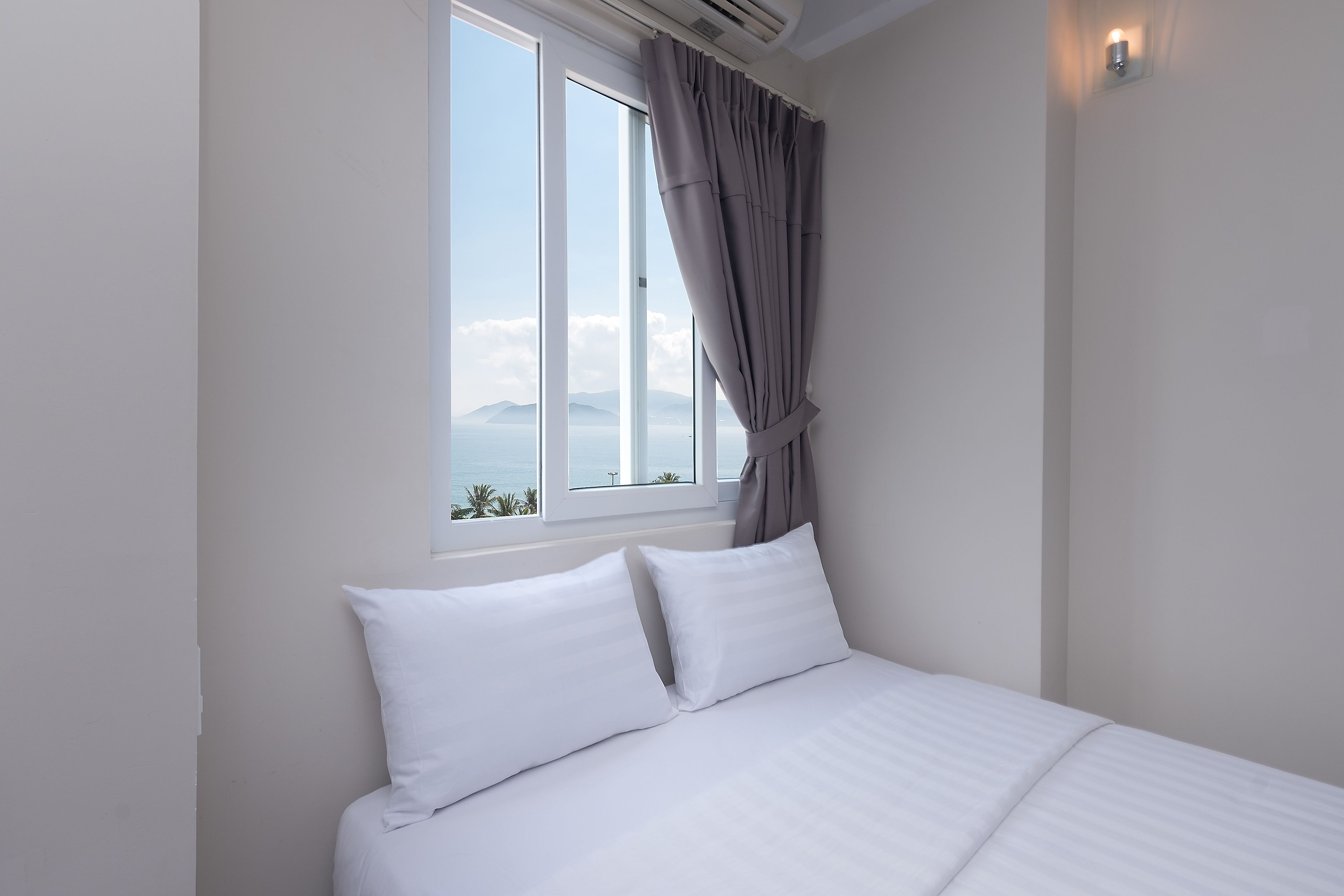 Quarto com cama, almofadas brancas, janela com cortinas e vista para o mar e montanhas.