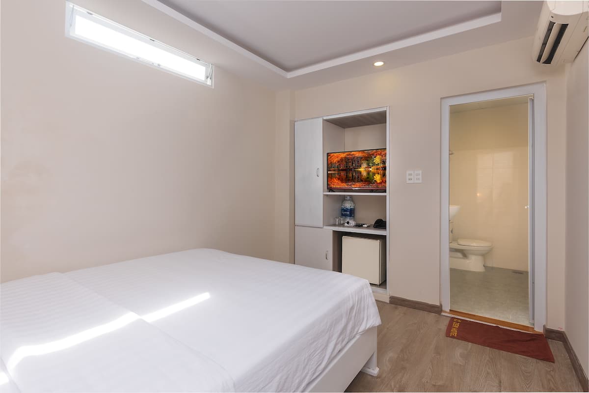 Quarto de hotel com cama, guarda-roupa, secretária pequena com garrafa de água e banheiro visível por uma porta aberta.