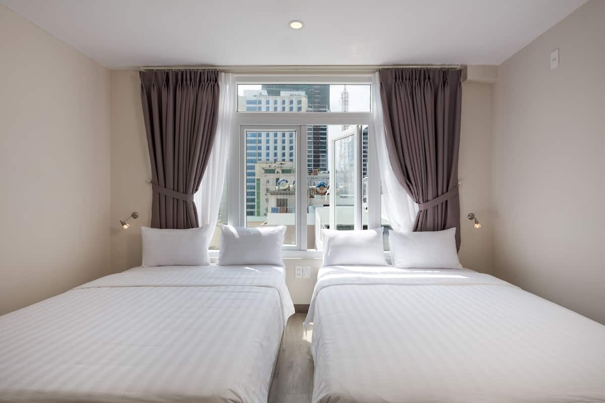 Quarto de hotel com duas camas, uma janela grande com cortinas e vista para edifícios.
