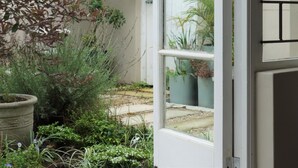 Une porte ouverte donnant sur un jardin où l’on peut voir différentes plantes et un mur blanc.