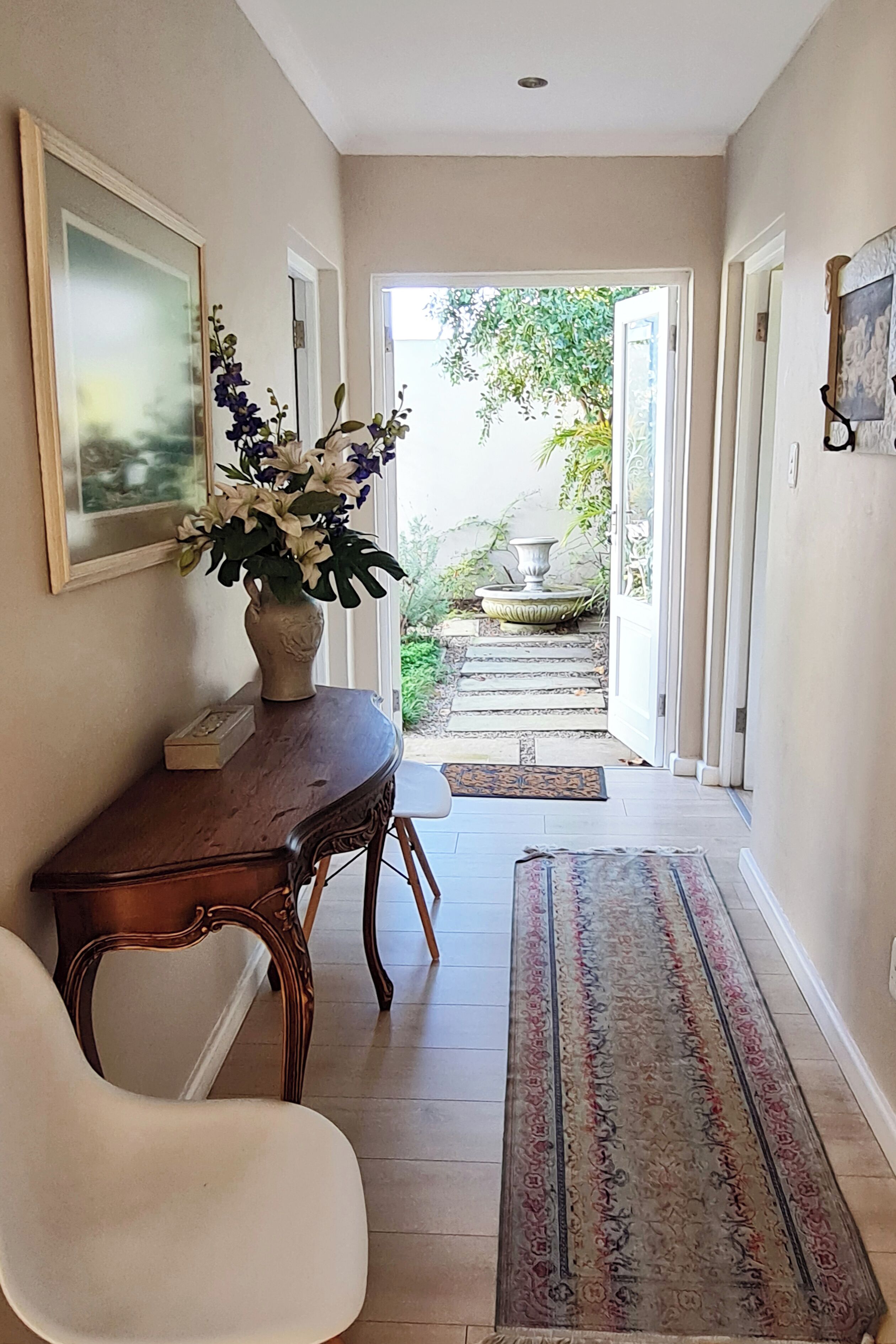Un couloir avec une table en bois, un vase de fleurs, un tableau encadré et un tapis menant à un espace extérieur doté de marches et d’un jardinière.