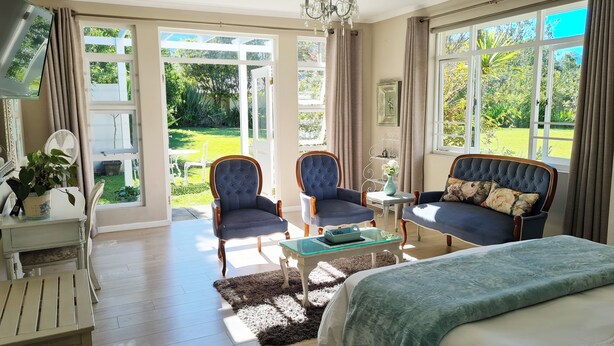 Une chambre spacieuse avec un grand lit, deux fauteuils, une petite table et une vue sur un jardin grâce à de grandes fenêtres.