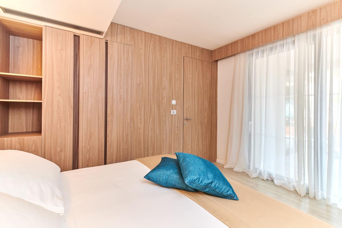 Une chambre à coucher moderne avec une armoire en bois, un lit agrémenté d’oreillers blancs et bleus, et des rideaux légers.