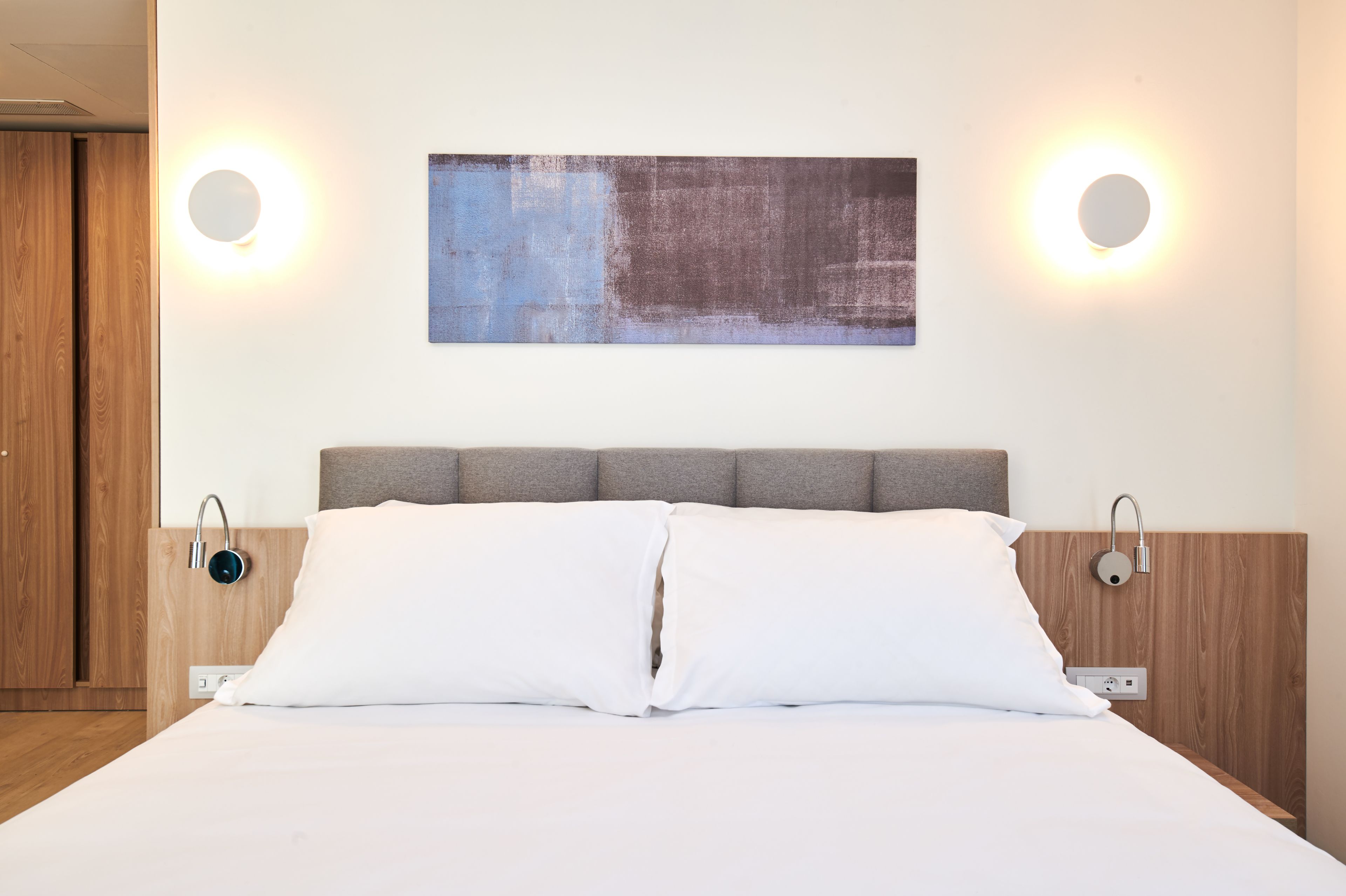 Una habitación de hotel moderna con una cama grande, dos mesitas de noche, un cabecero y una obra de arte colgada en la pared.