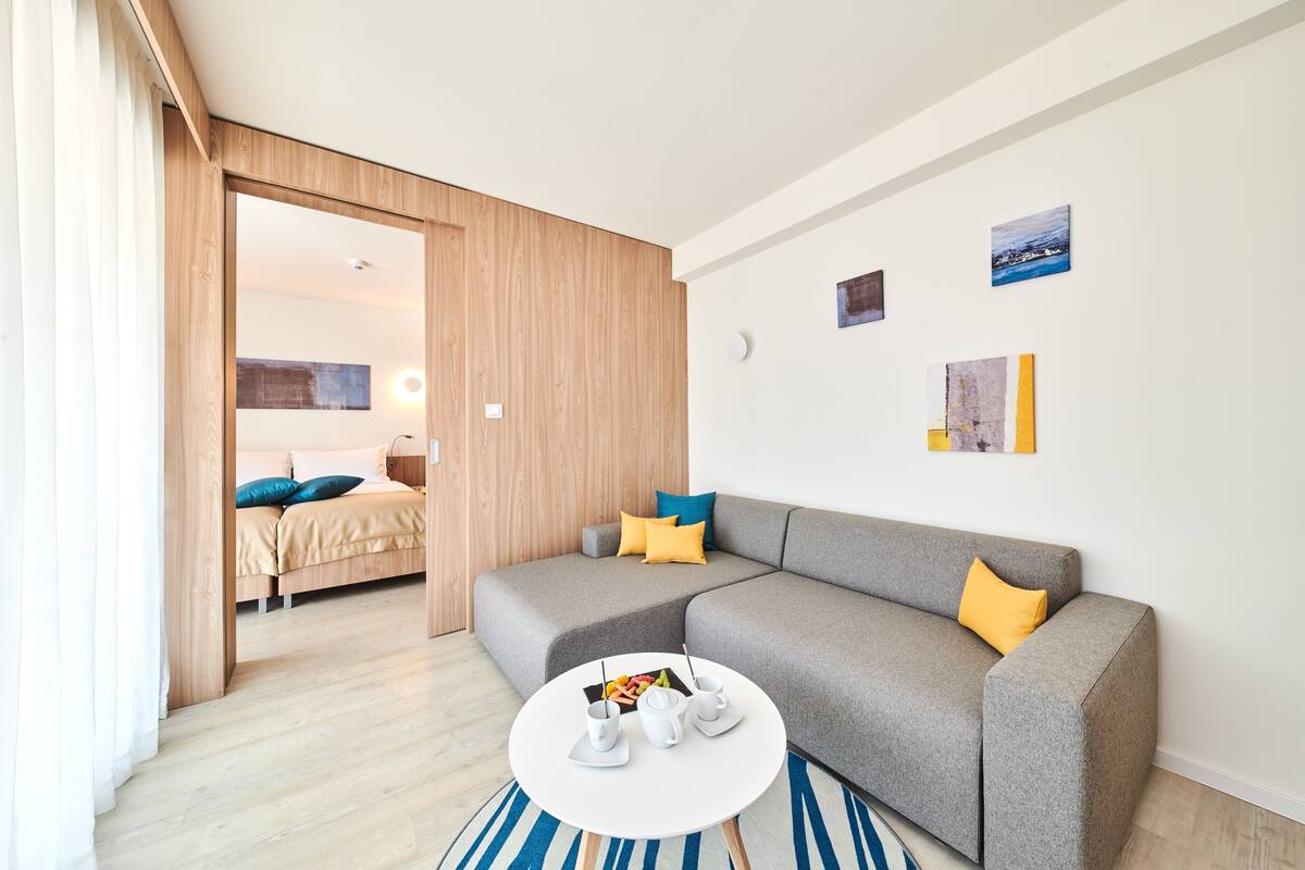 Un salon moderne avec un canapé gris, une table basse ronde et un lit à l’arrière-plan.
