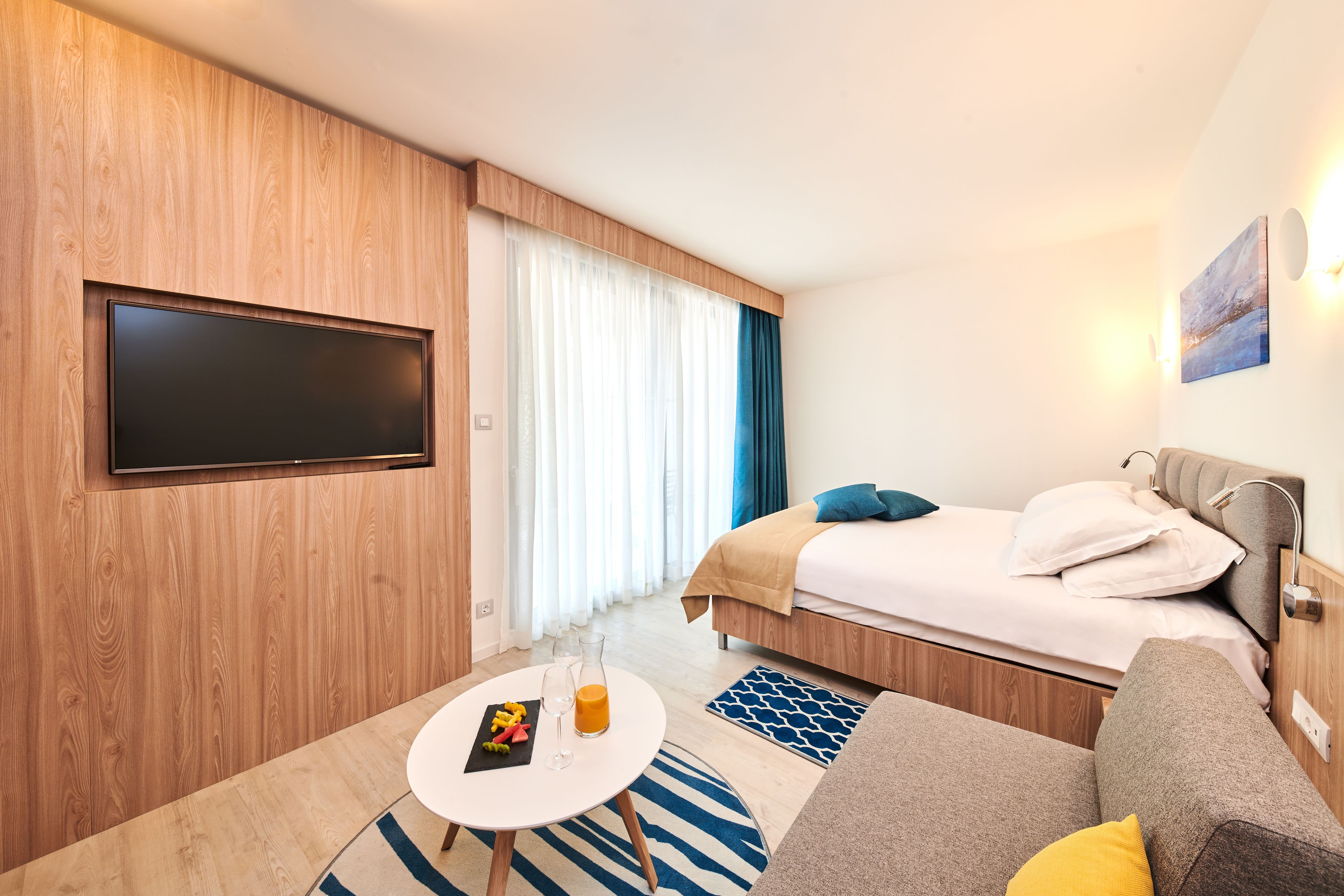 A modern hotel room with a large flat-screen TV mounted on a wooden wall, a bed with a headboard, a small round table with a glass of orange juice and a fruit plate, and a sofa with a yellow pillow.