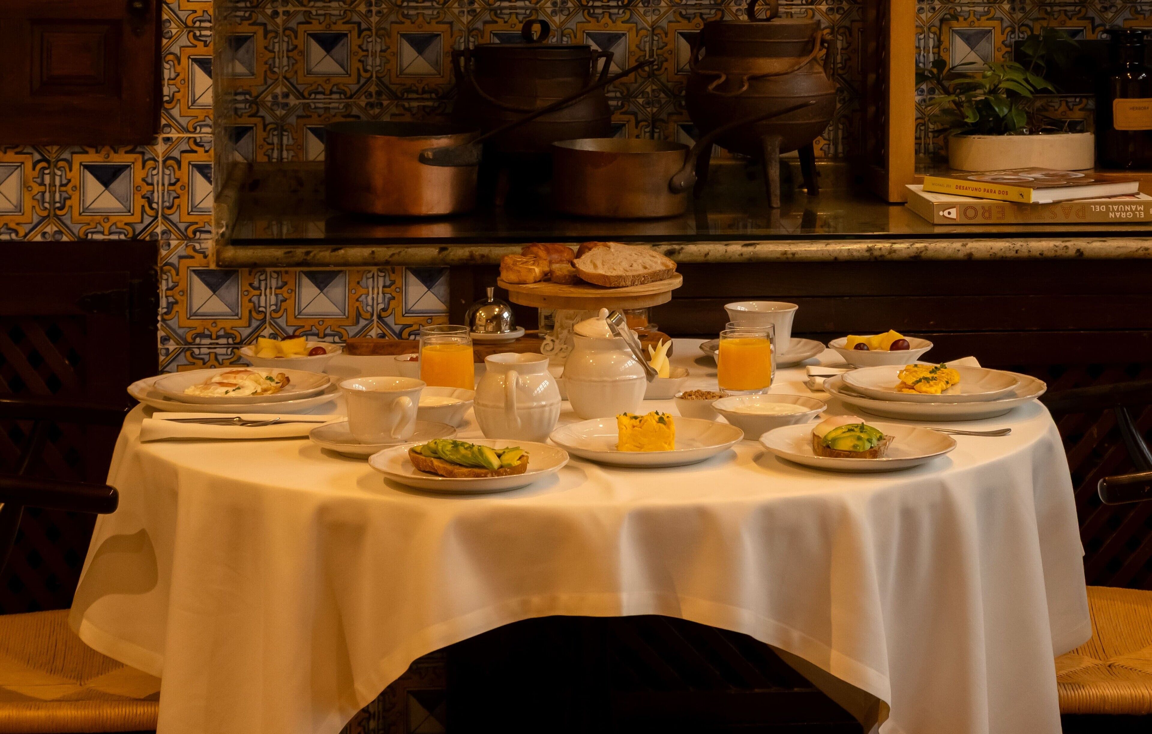 A breakfast table set with various dishes, including toast, fruit, and beverages, in a room with patterned tile walls and kitchen utensils in the background.