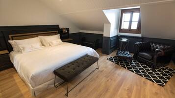 A bedroom with a large bed, a bench, a leather armchair, a small table, and a window with natural light.