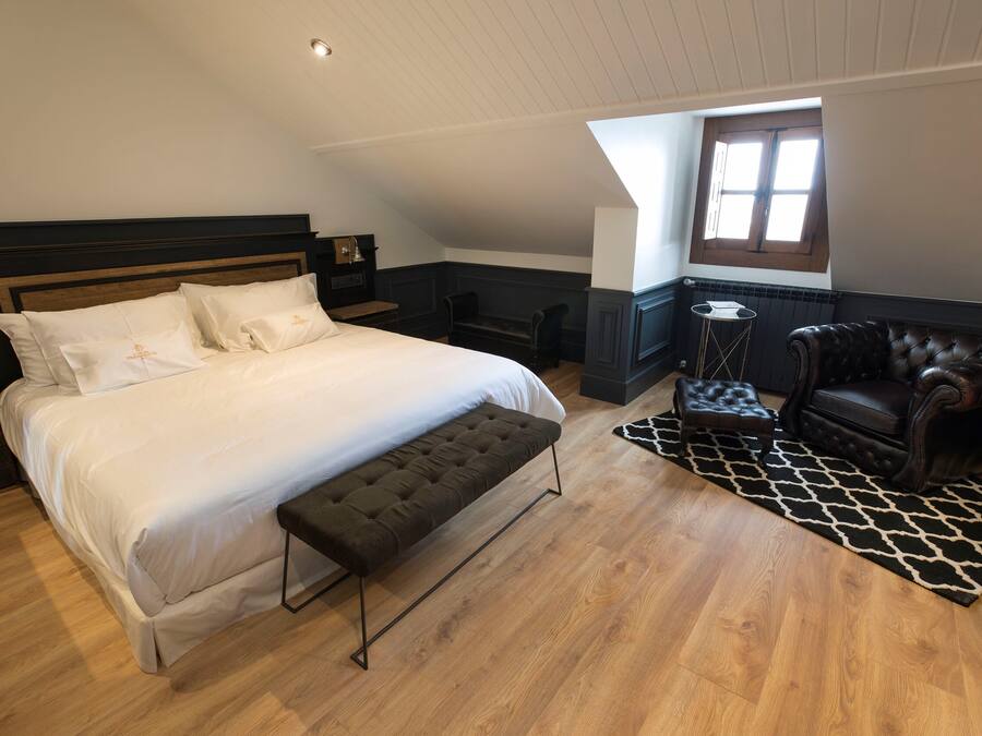 A bedroom with a large bed, a bench, a leather armchair, a small table, and a window with natural light.