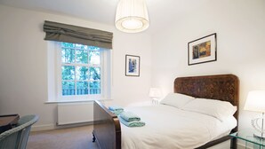 Um quarto bem arrumado com uma cama grande, uma janela com cortinas, uma mesa de cabeceira com abajur e uma cadeira.
