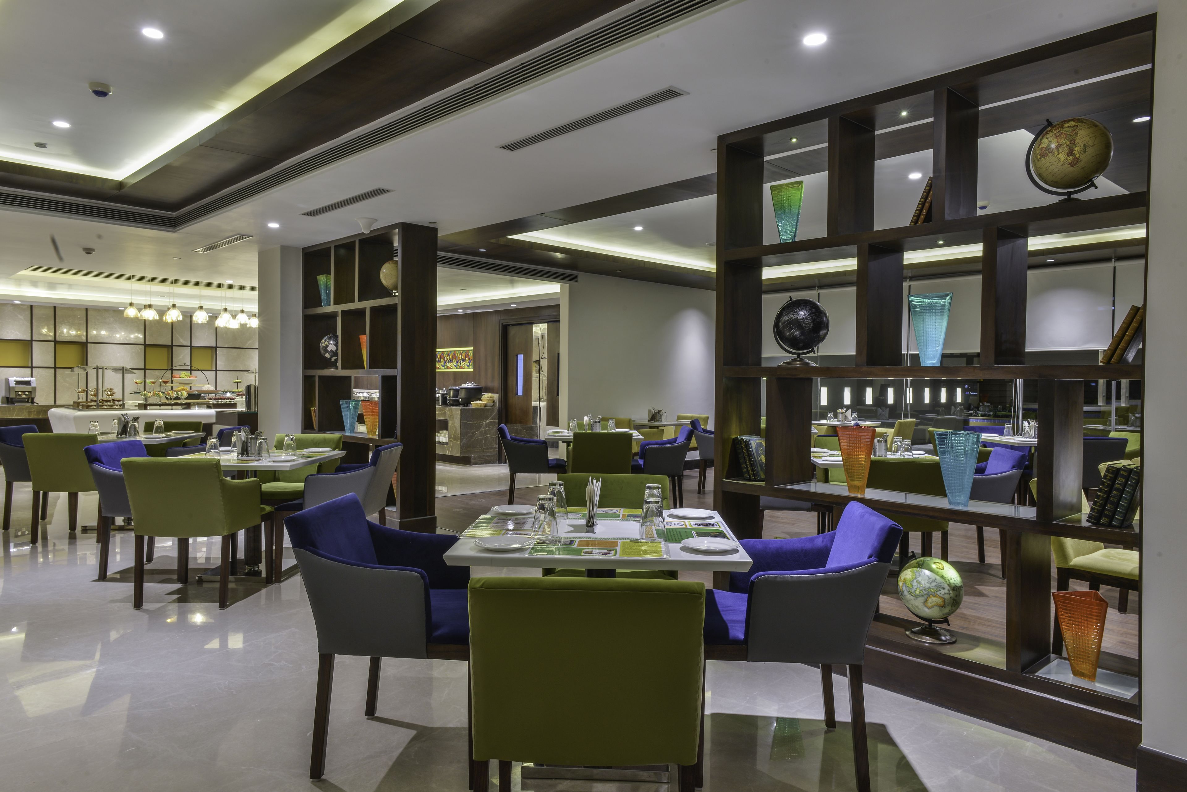 A modern dining area with green and blue chairs, tables set with white plates and silverware, and a decorative globe on a shelf.