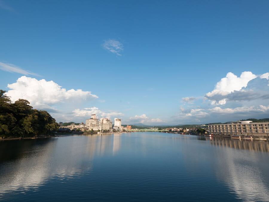 A calm river with buildings on the banks, including a large hotel with multiple floors and a smaller building nearby.