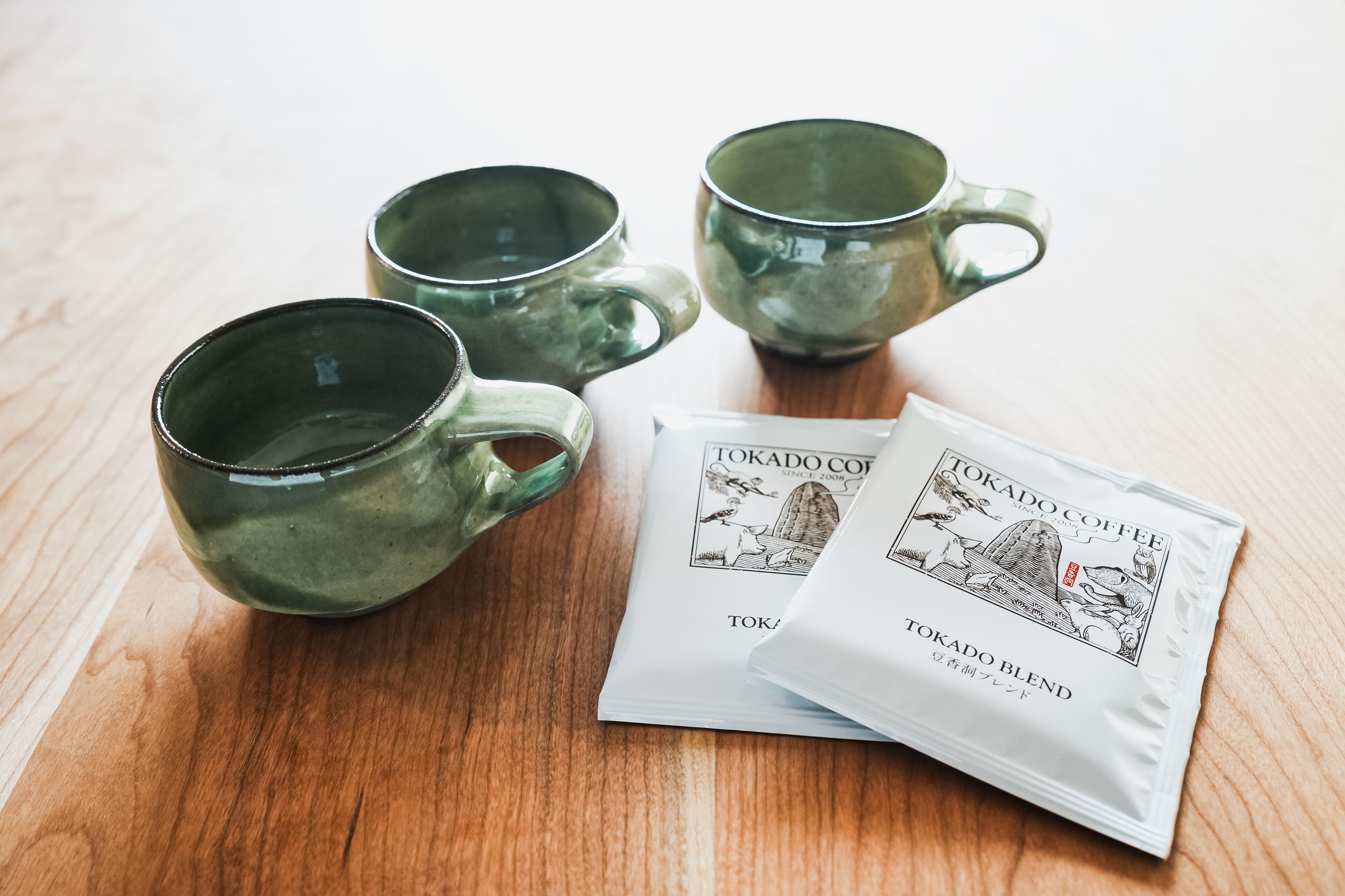 Neljä vihreää keramiikkakuppia ja kaksi pakettia Tokado-kahvisekoitusta puupinnalla.