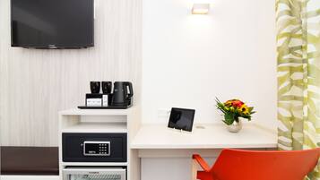 Habitación de hotel con televisión de pantalla plana, cafetera, tableta y un florero sobre un escritorio blanco.
