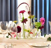 Mesa posta para refeição, com vários vasos de flores e taças de vinho.