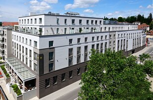 Um edifício moderno de vários andares com telhado plano, janelas amplas e uma área externa coberta com assentos.