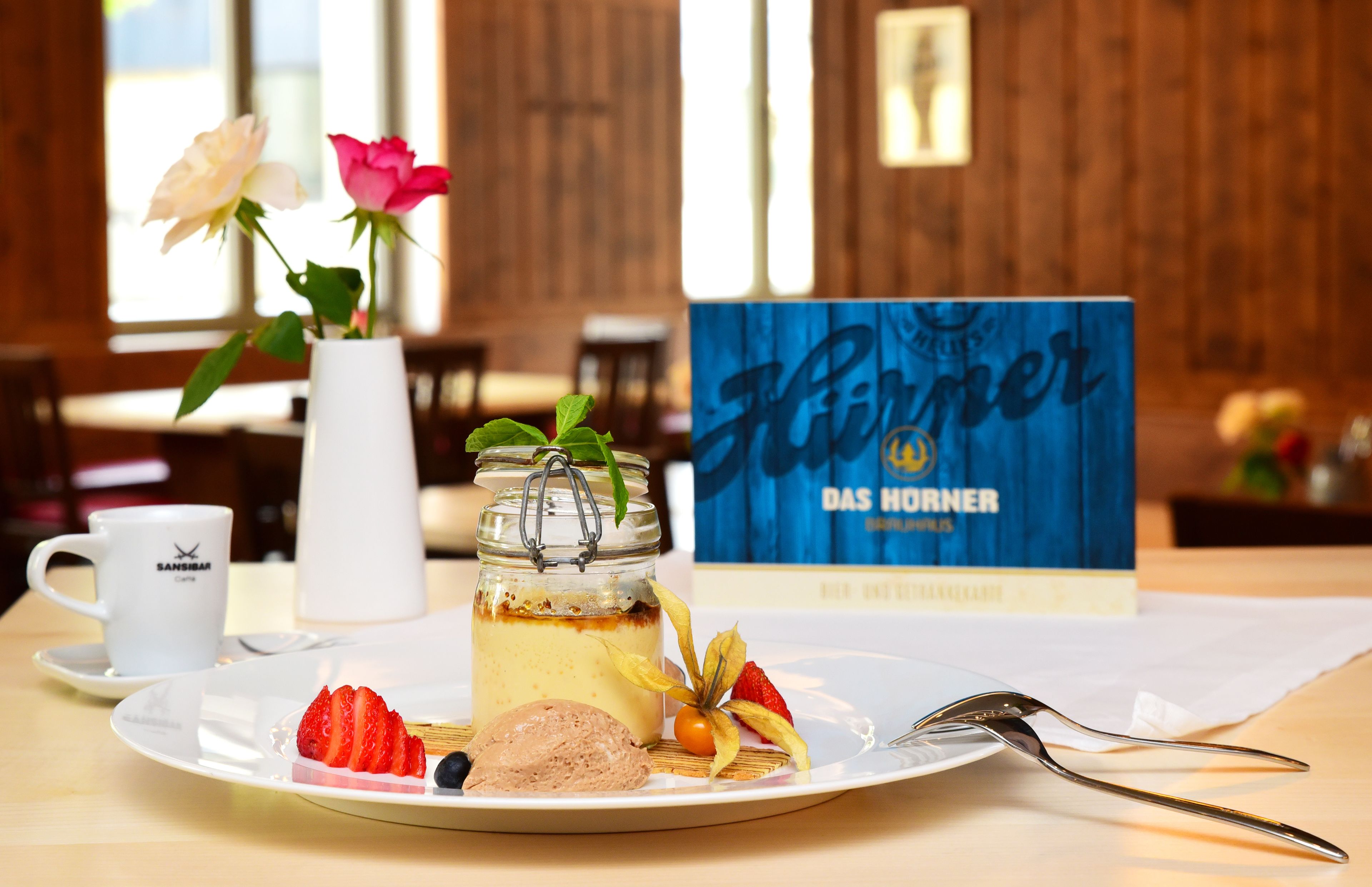 A dessert plate with strawberries, blueberries, and a scoop of ice cream, a white cup, and a vase with flowers on a table in a room with wooden walls and a window.