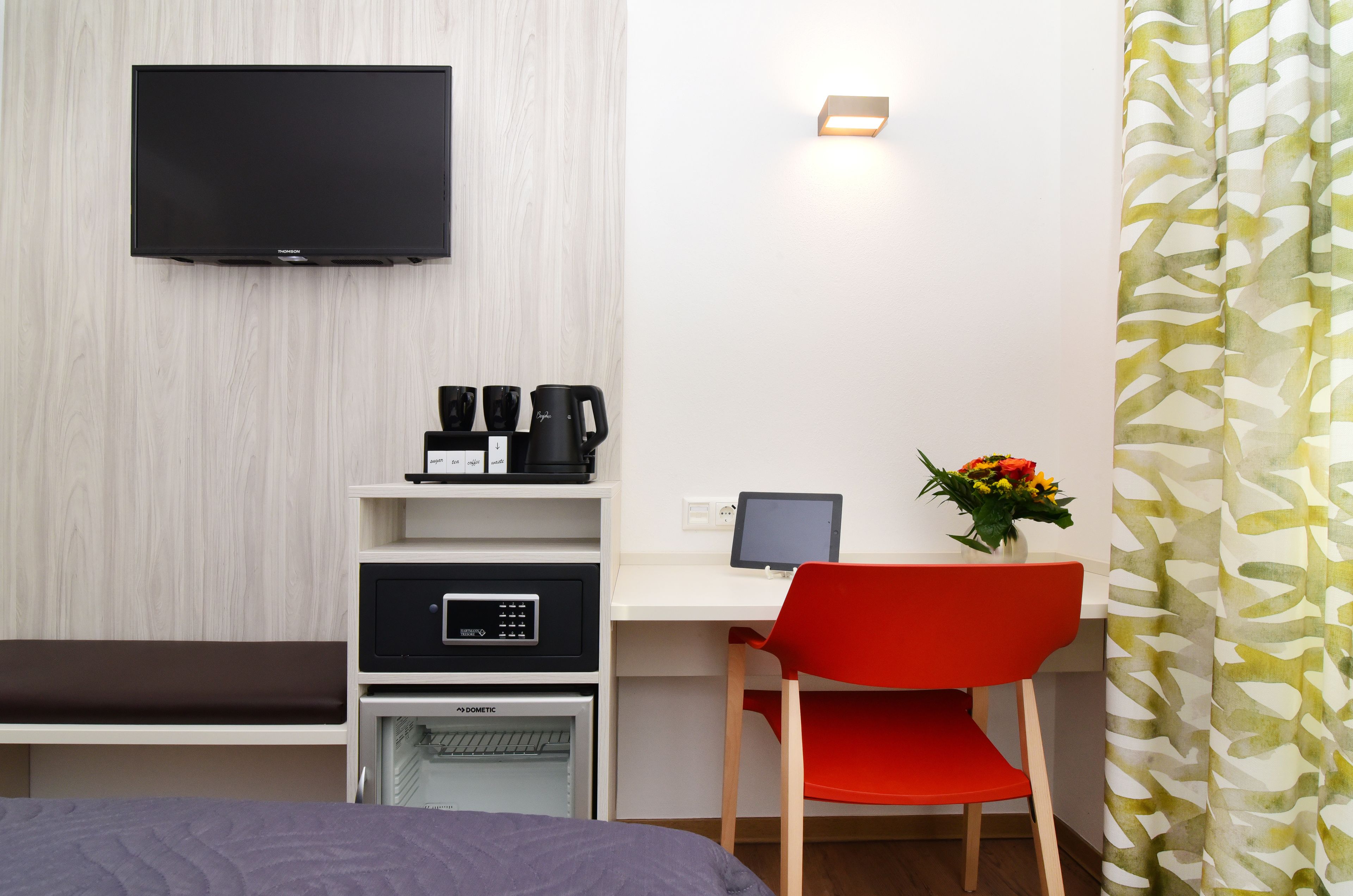 Habitación de hotel con televisión de pantalla plana en la pared, cafetera, un ordenador portátil sobre un escritorio, una silla roja y una cama con colcha morada.