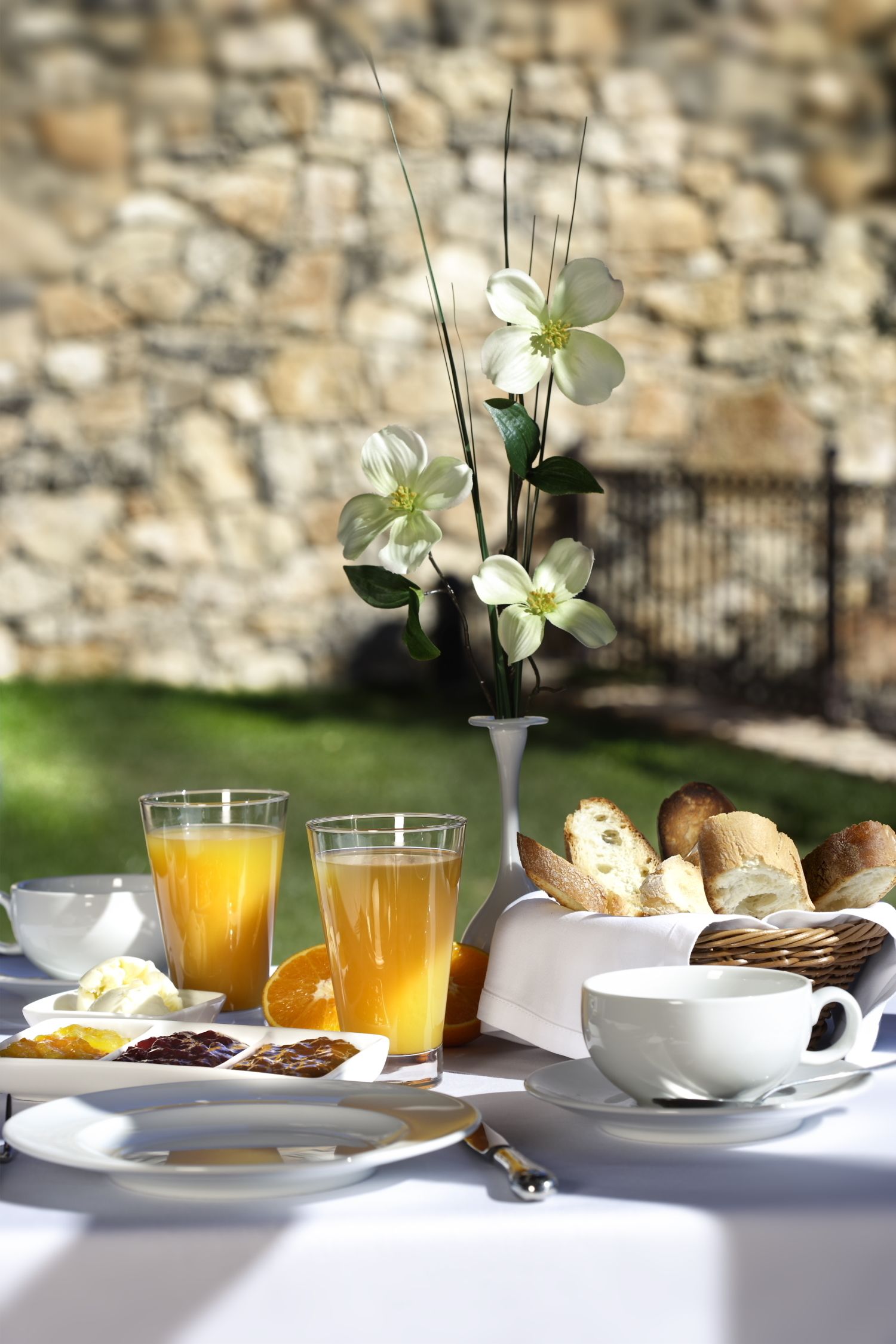 En frukostuppdukning med en stenmur i bakgrunden, en vit vas med blommor, två glas apelsinjuice, en kopp, en tallrik med bröd och en liten skål med frukt.