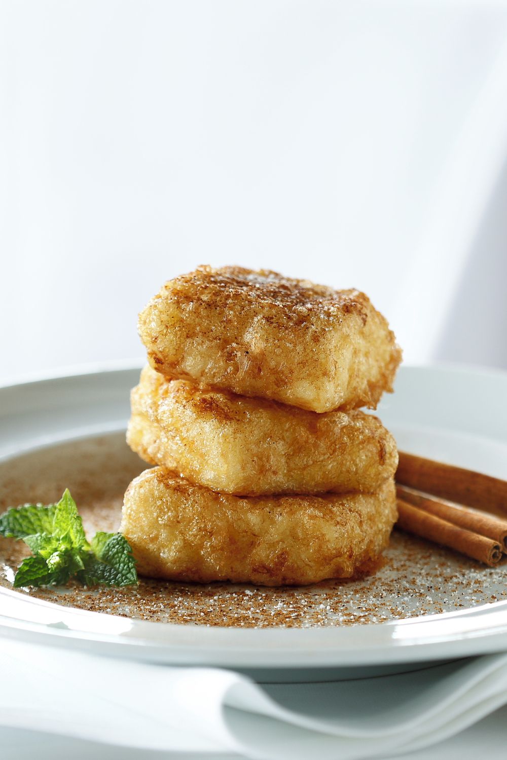 A stack of round, golden-brown pastries on a white plate with a dusting of cinnamon and a mint leaf garnish.