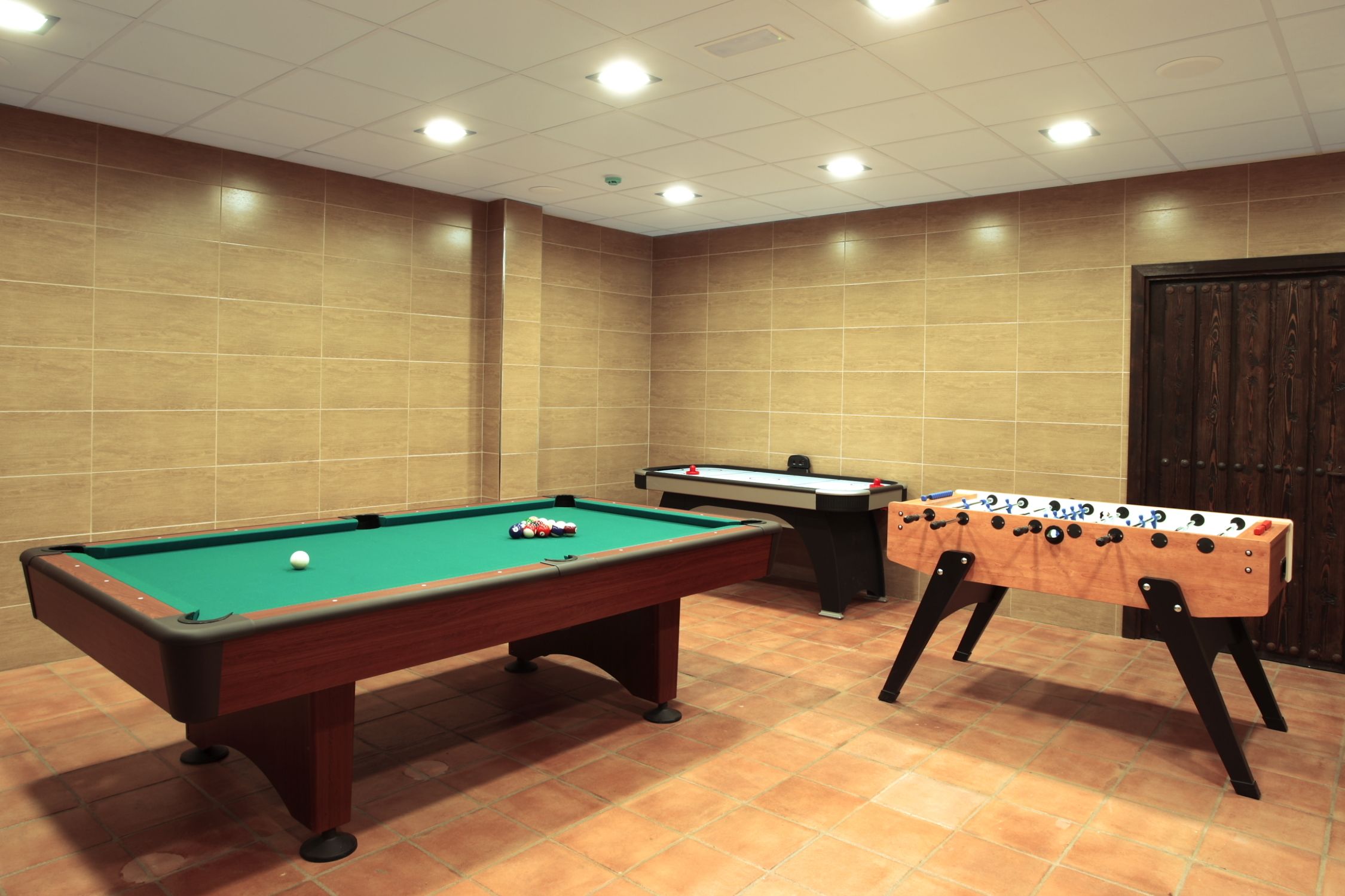 A pool table, a foosball table, and a billiards table in a room with tiled walls and a wooden door.