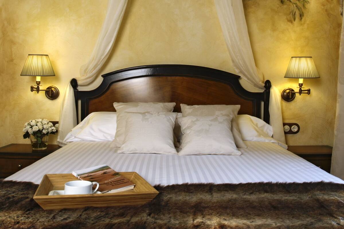 A bedroom with a canopy bed, two bedside lamps, a nightstand with a vase of flowers, and a tray with a book and a cup.