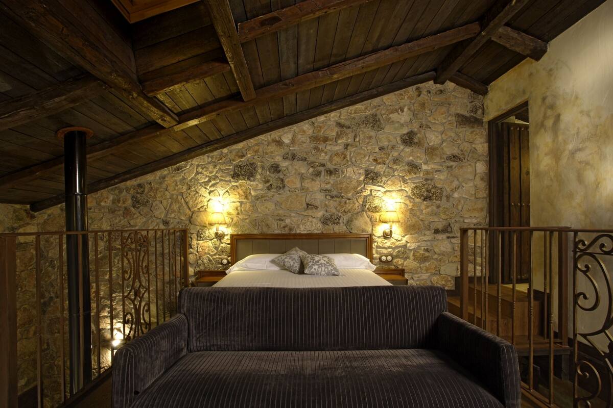 A bedroom with a stone wall, wooden beams, a bed with pillows, and two wall-mounted lights.