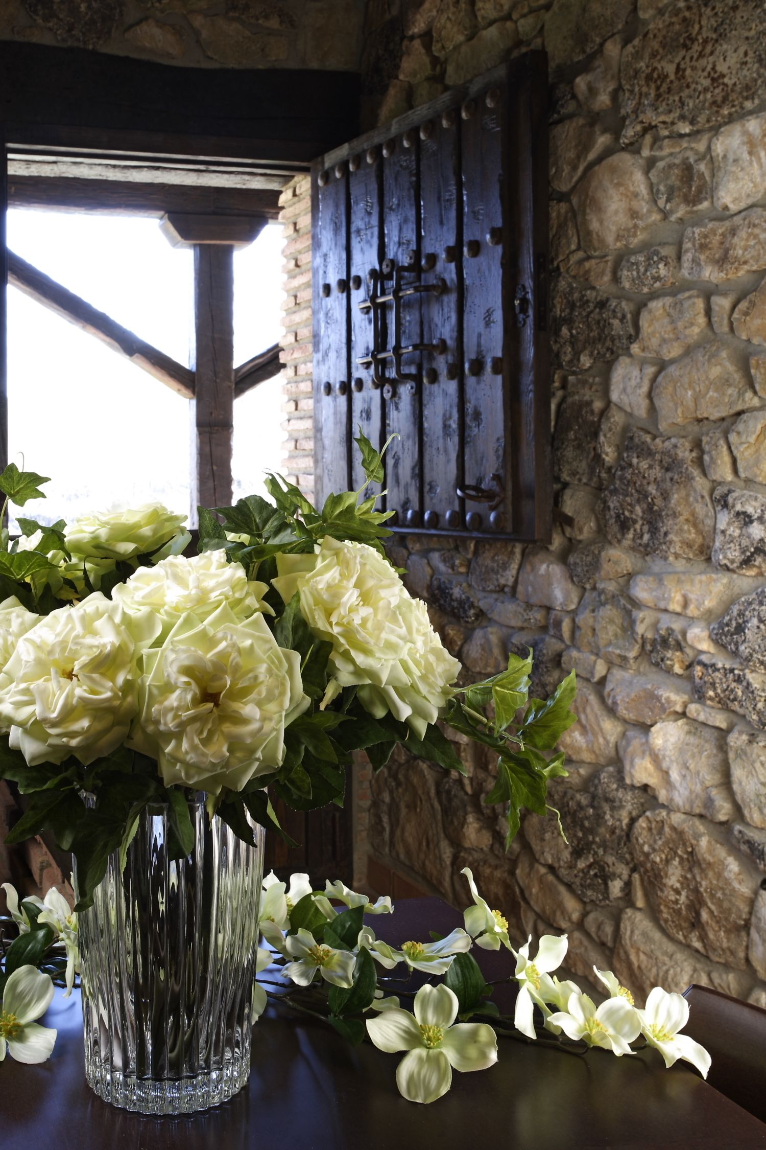 A table with a clear glass vase containing white flowers, set against a stone wall with a rustic wooden door.