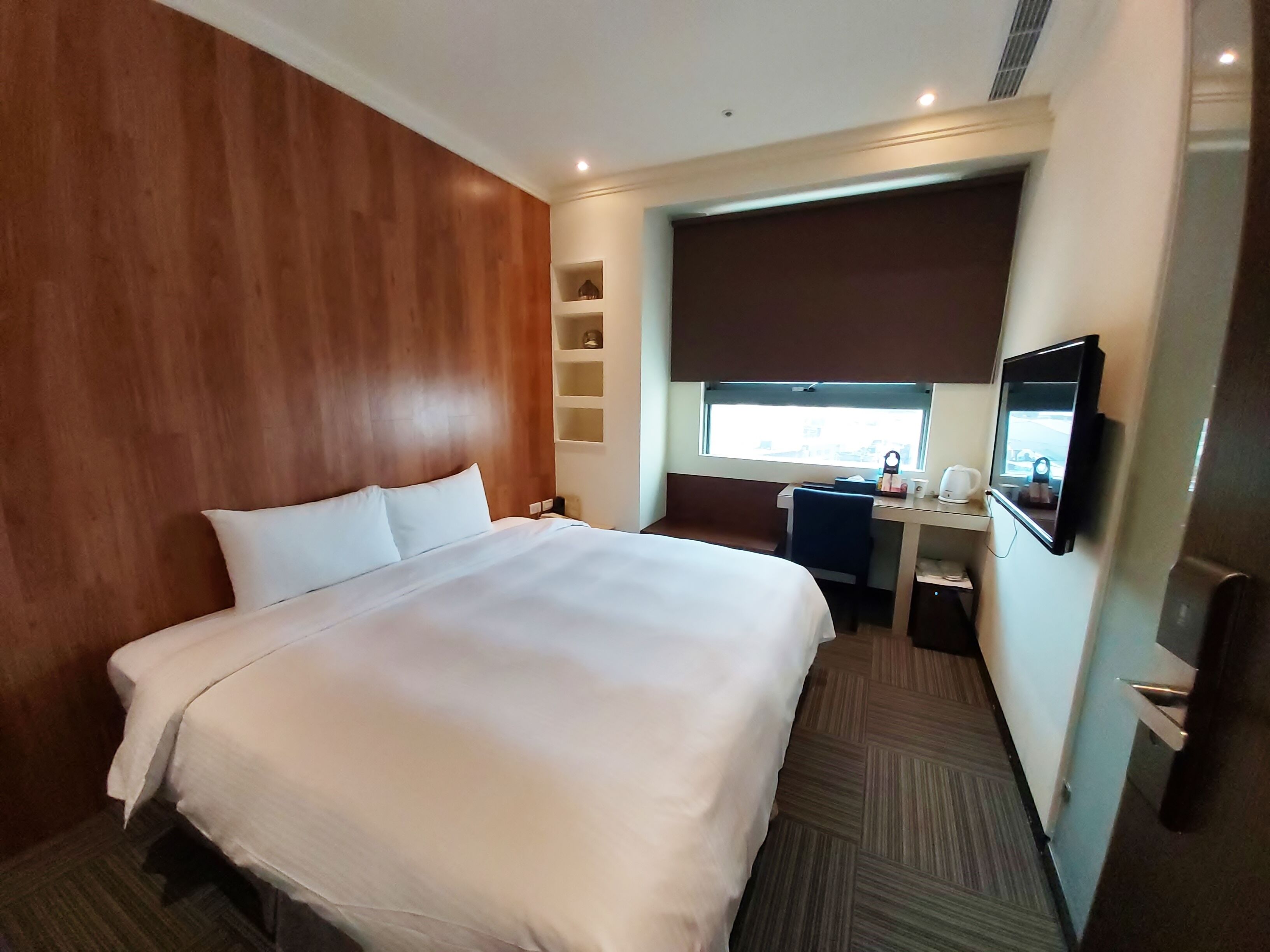 A hotel room with a large bed, a desk, a television, and a window with a view of the city.