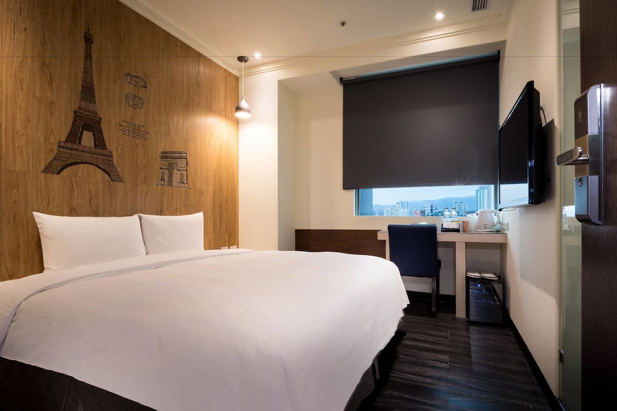 A hotel room with a large bed, a desk with a chair, a television, and a window with a city view.