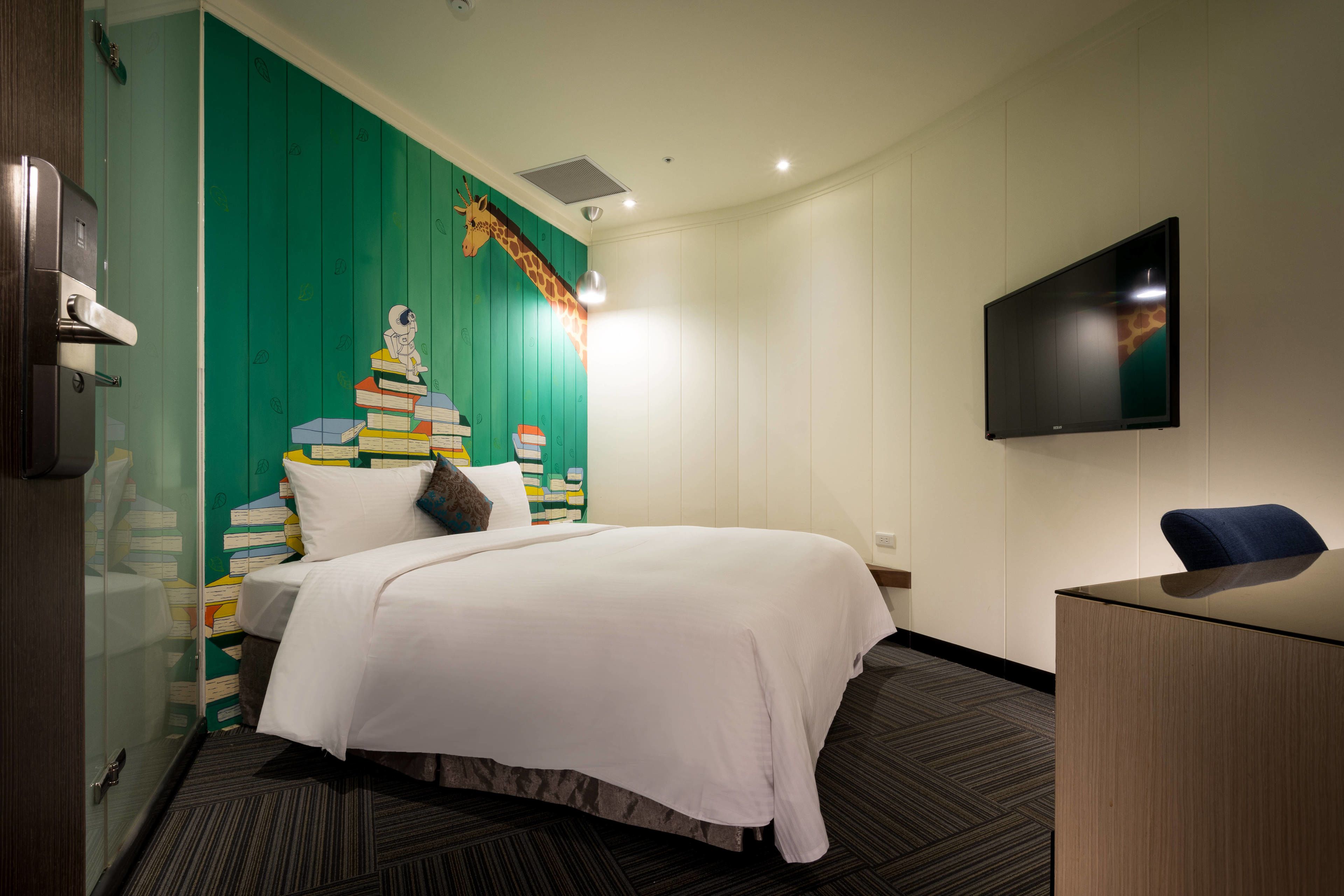 A hotel room with a bed, a desk, a TV, and a wall with a giraffe and books.