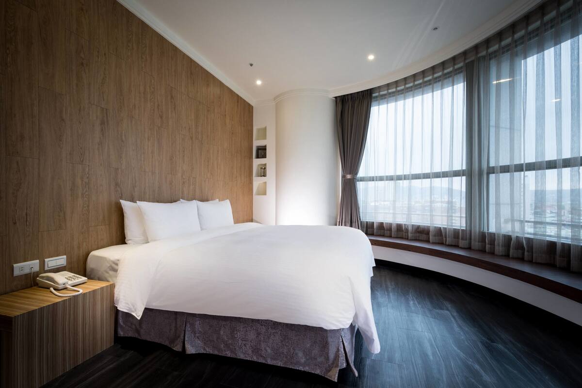 A modern hotel room with a large bed, wooden paneling on one wall, a bedside telephone, and a large window with curtains.