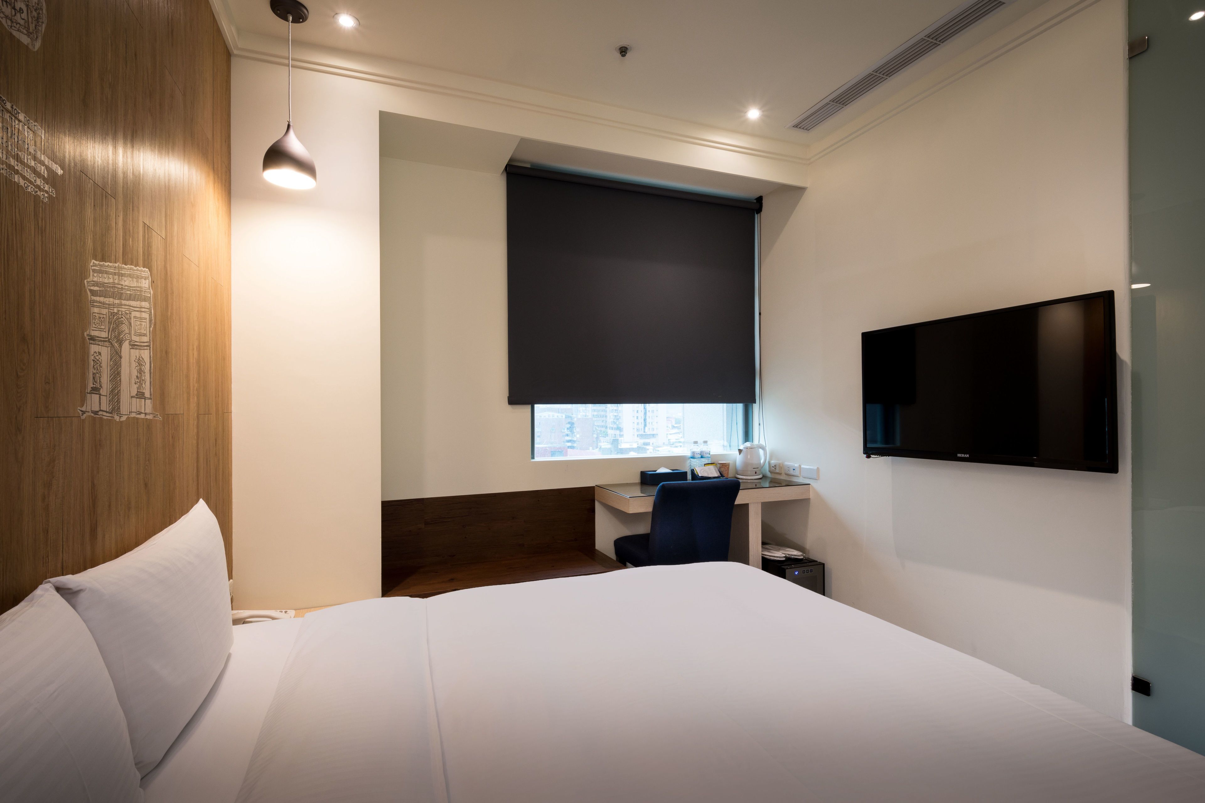 A hotel room with a large bed, a desk with a chair, a flat-screen TV, a window with a blind, and a wooden accent wall.