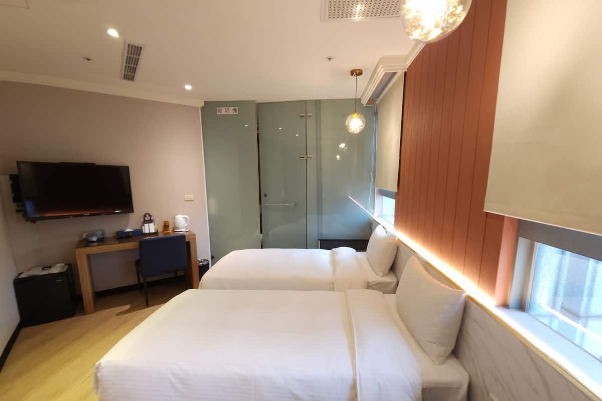 A hotel room with two beds, a desk with a chair, a television, and a window with blinds.