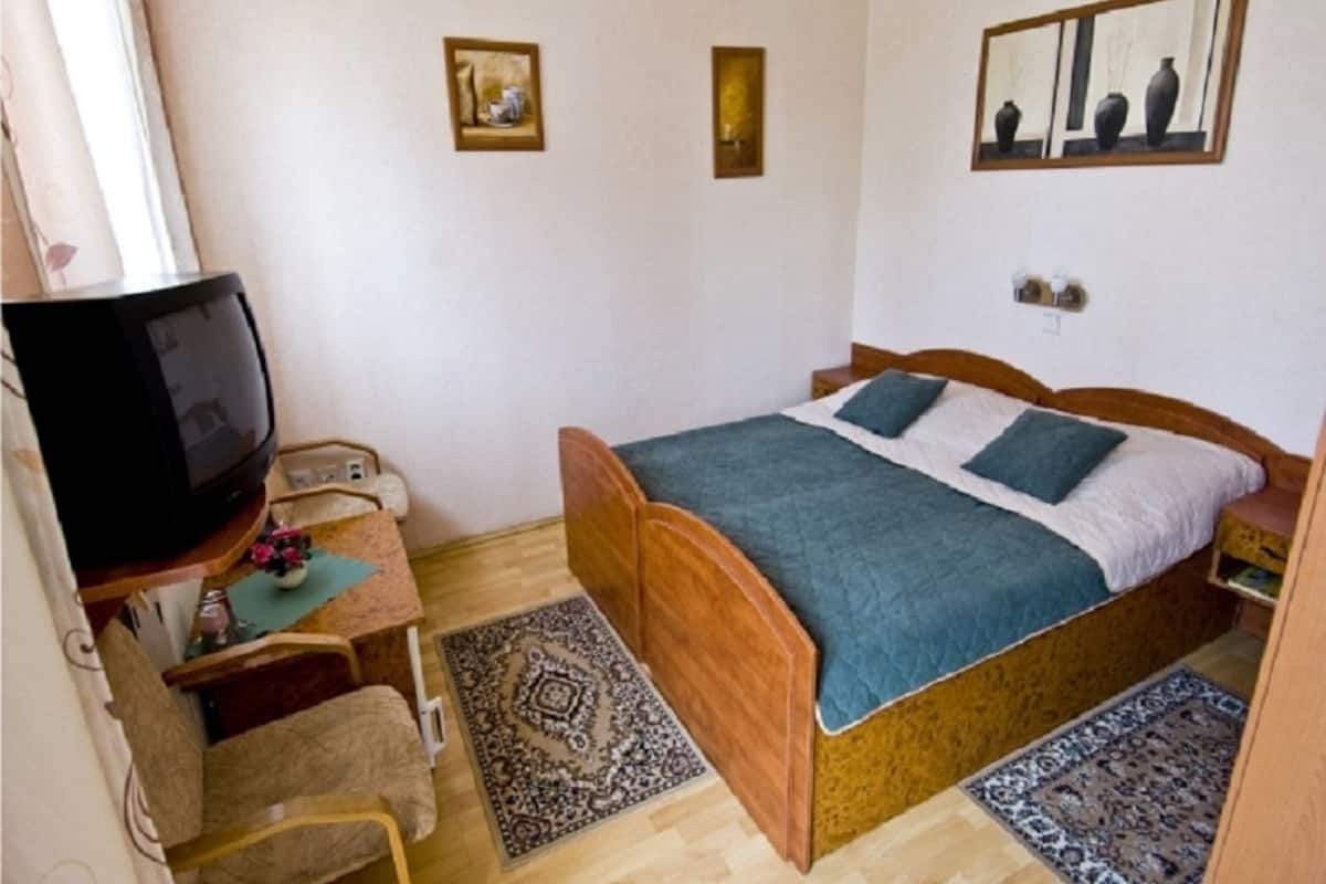 Ein Schlafzimmer mit einem hölzernen Bett, einem Fernseher auf einem Schrank, einem Schreibtisch mit Stuhl und zwei Teppichen auf dem Boden.