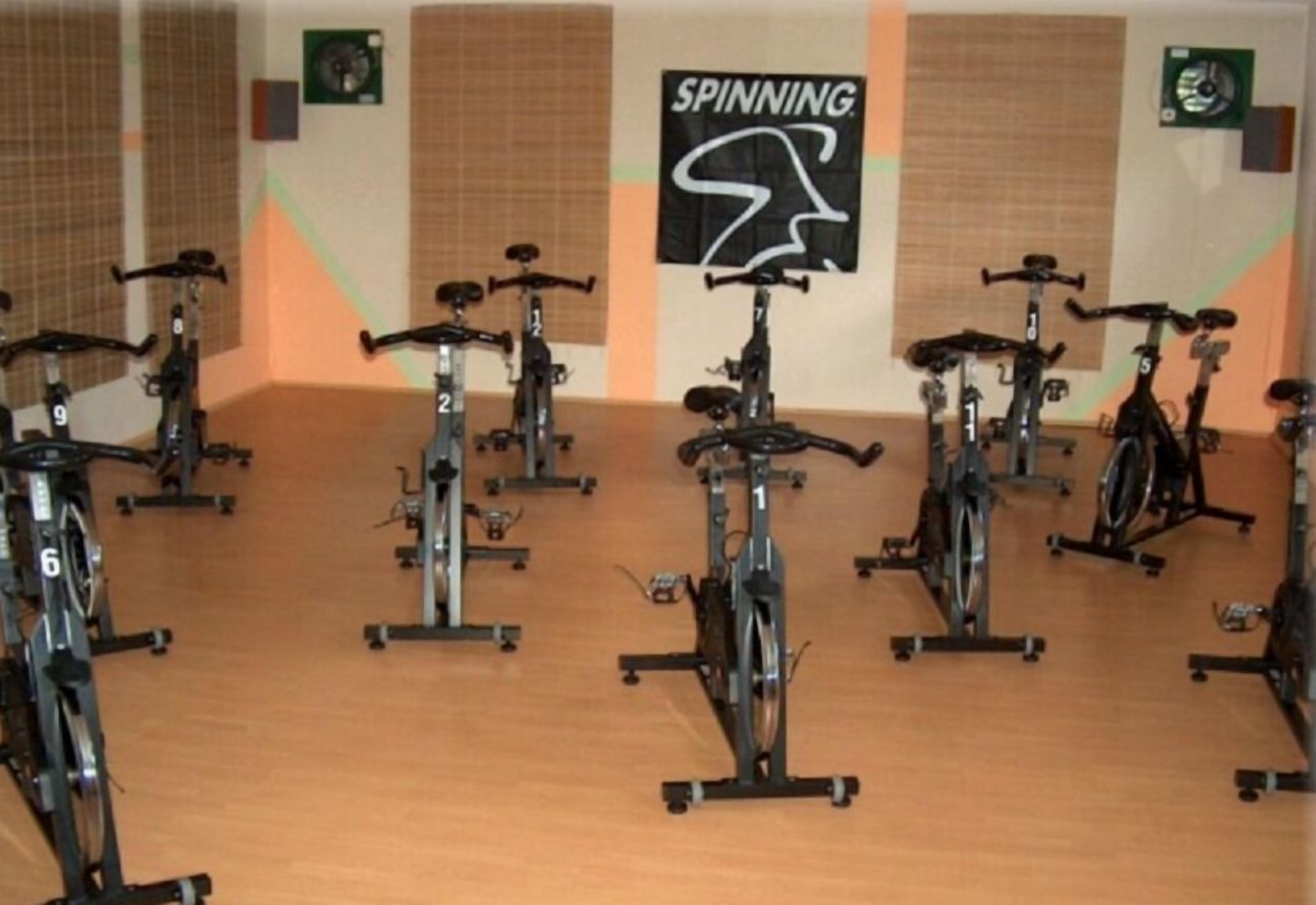A gym with stationary bikes arranged in rows, a sign that reads "SPINNING," and a fan on the wall.